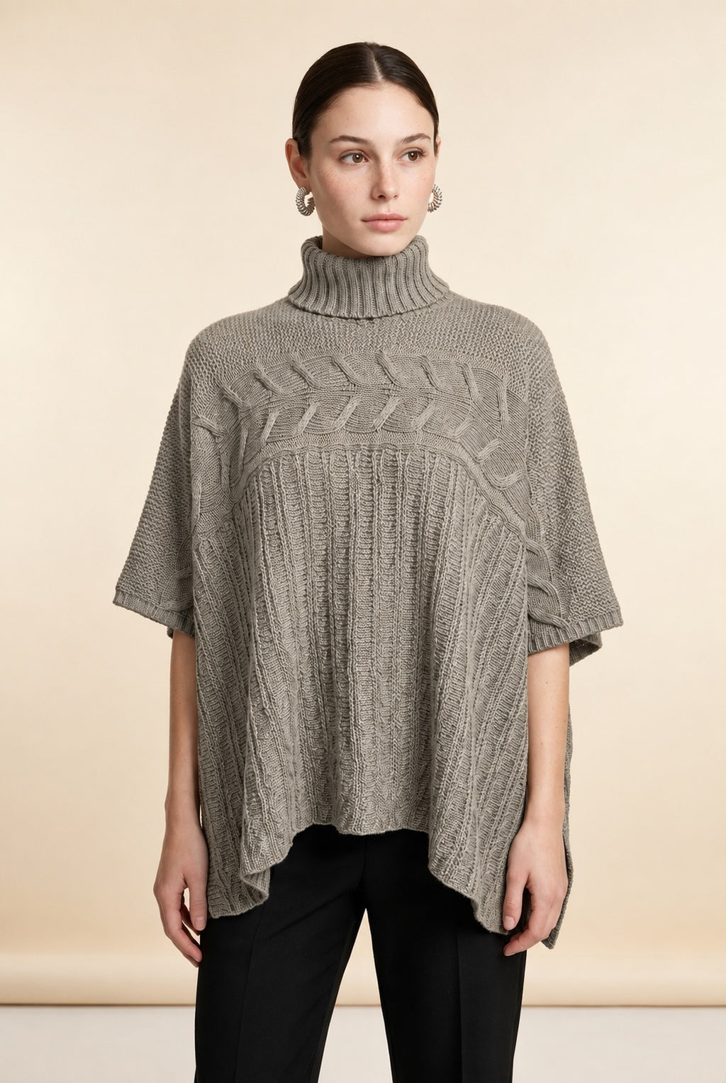 A woman wearing the Aosta cable-knit turtleneck sweater in taupe, highlighting its ribbed lower body and wide sleeves.