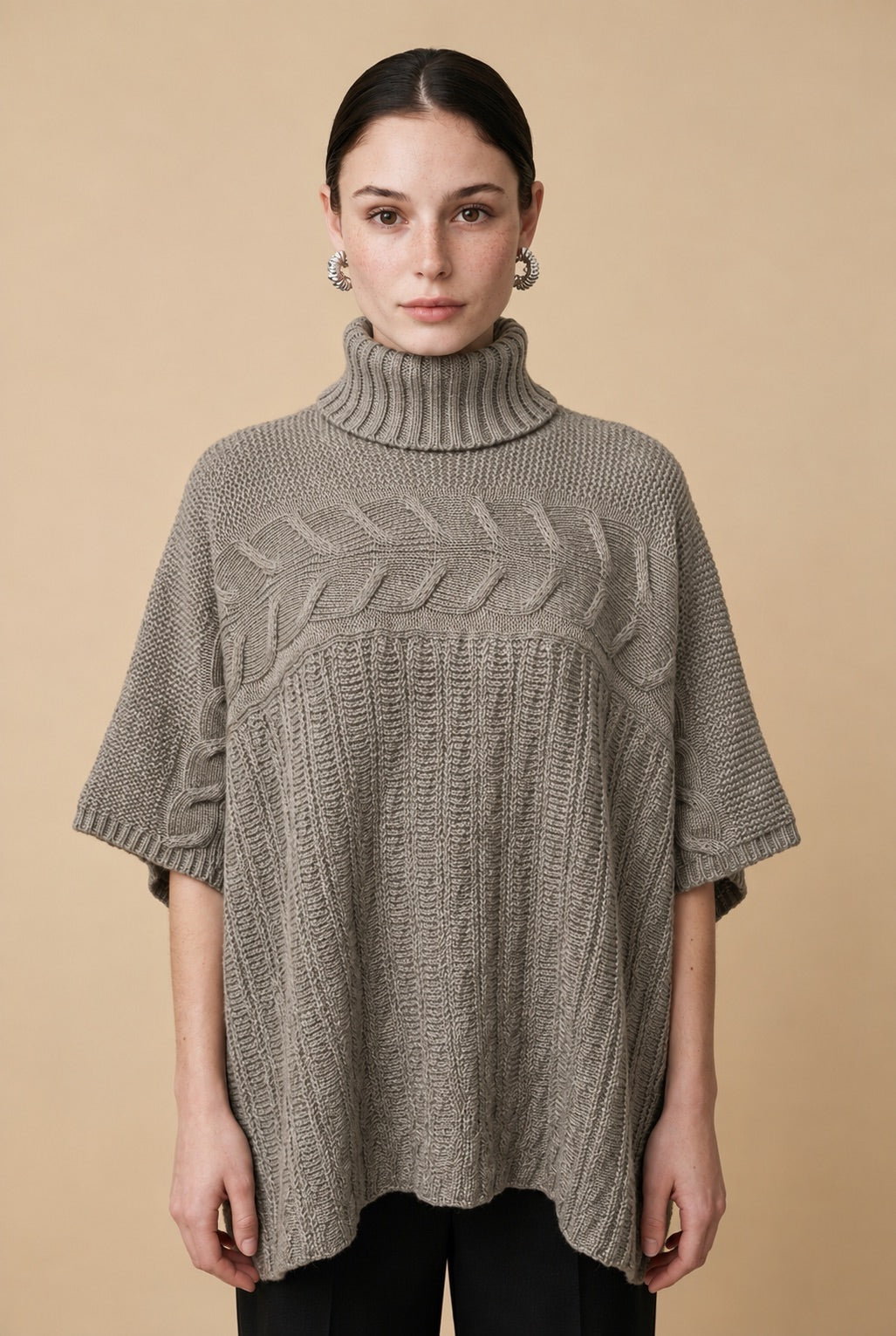 A woman wearing the Aosta cable-knit turtleneck sweater in taupe, highlighting its ribbed lower body and wide sleeves.
