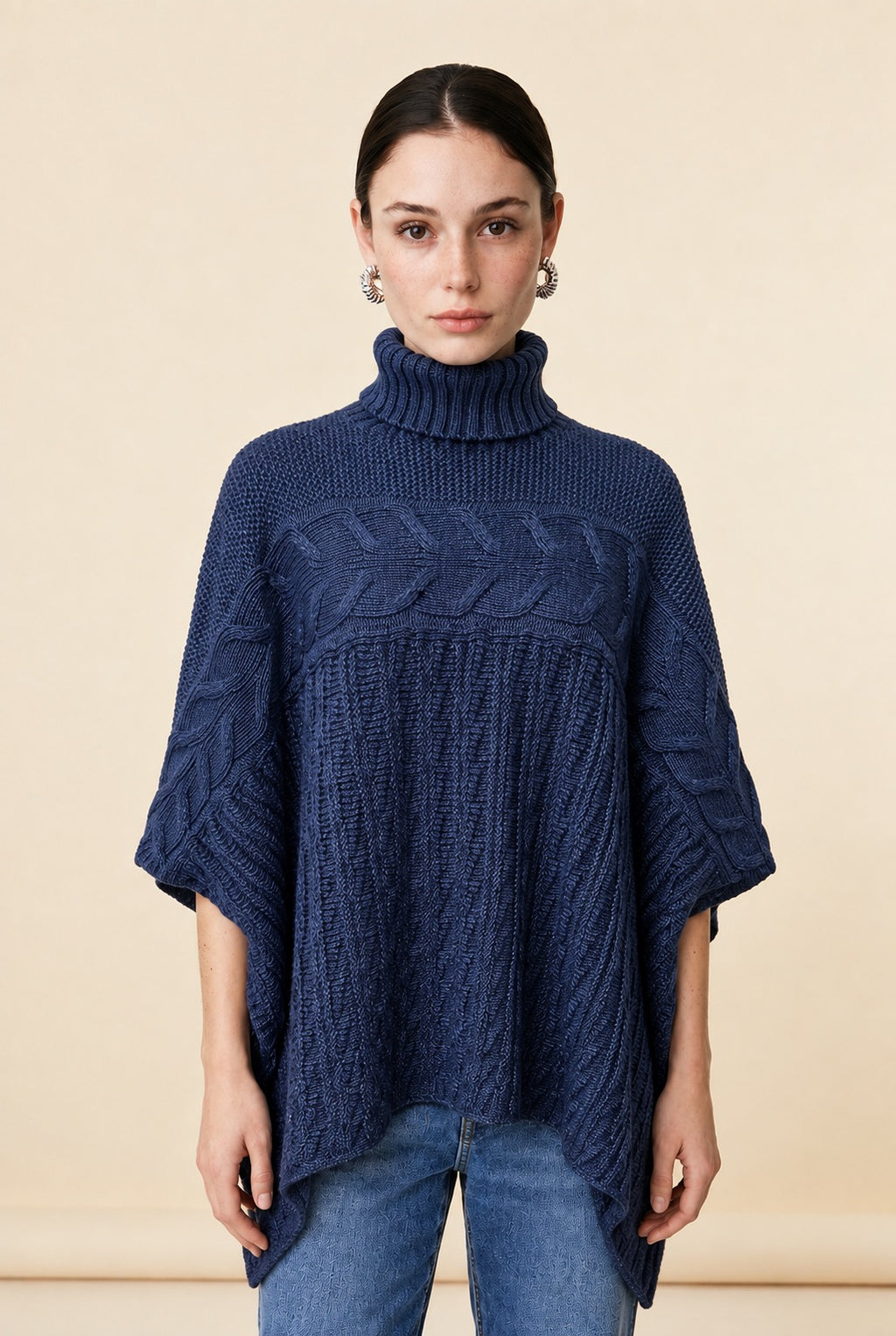 A woman wearing the Aosta cable-knit turtleneck sweater in navy blue, showcasing its poncho silhouette and textured weave.
