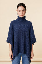 A woman wearing the Aosta cable-knit turtleneck sweater in navy blue, showcasing its poncho silhouette and textured weave.