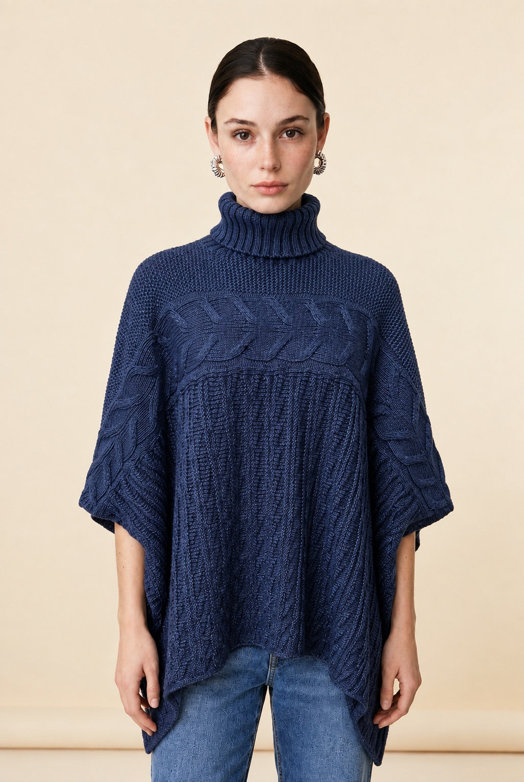 A woman wearing the Aosta cable-knit turtleneck sweater in navy blue, showcasing its poncho silhouette and textured weave.