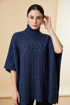 A woman wearing the Aosta cable-knit turtleneck sweater in navy blue, showcasing its poncho silhouette and textured weave.