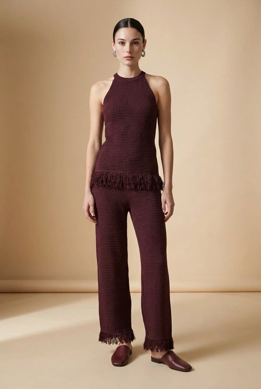 A woman wearing the Set Bérgamo knit two-piece set in deep burgundy, featuring a sleeveless halter top and straight-leg trousers with fringe hem details.
