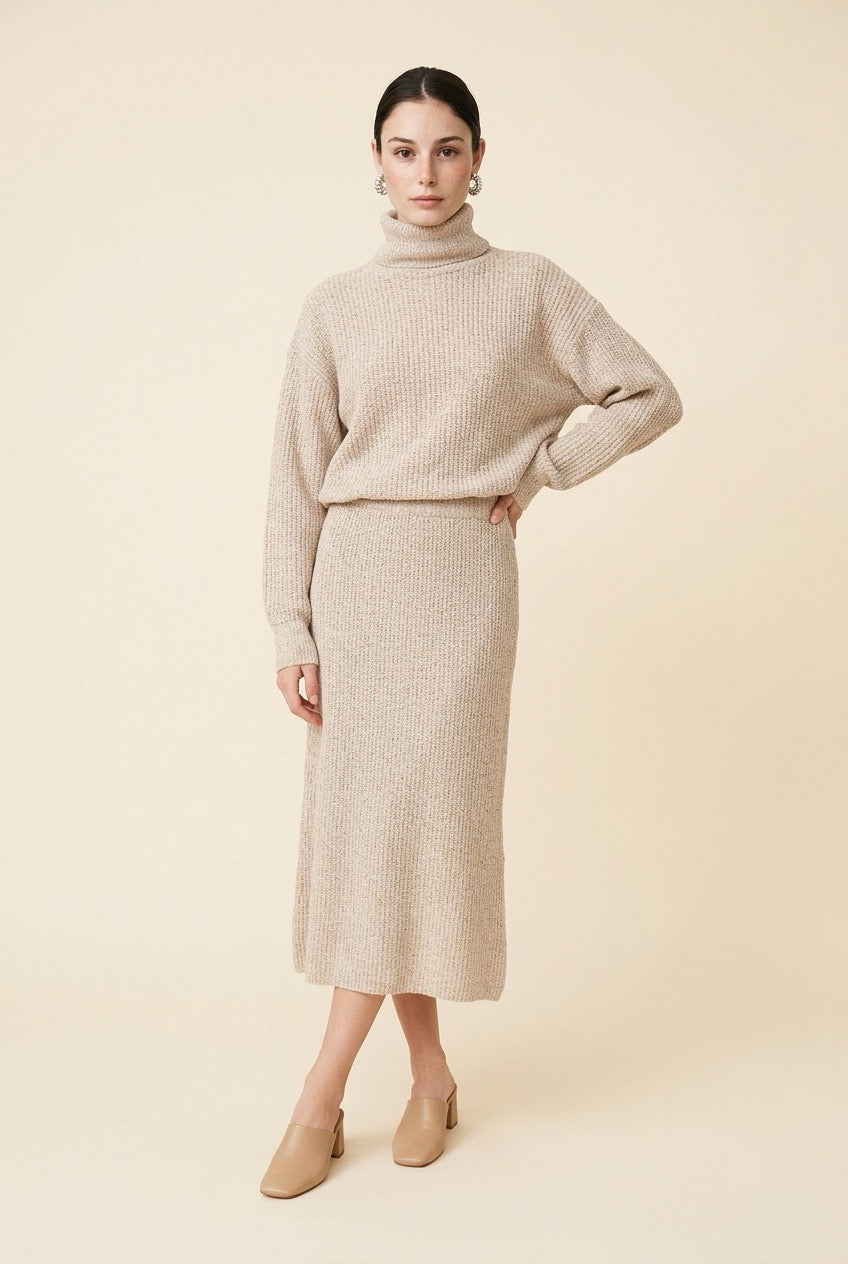 A woman wearing the Ginevra two-piece ribbed knit set in beige, featuring a turtleneck sweater and matching midi skirt.