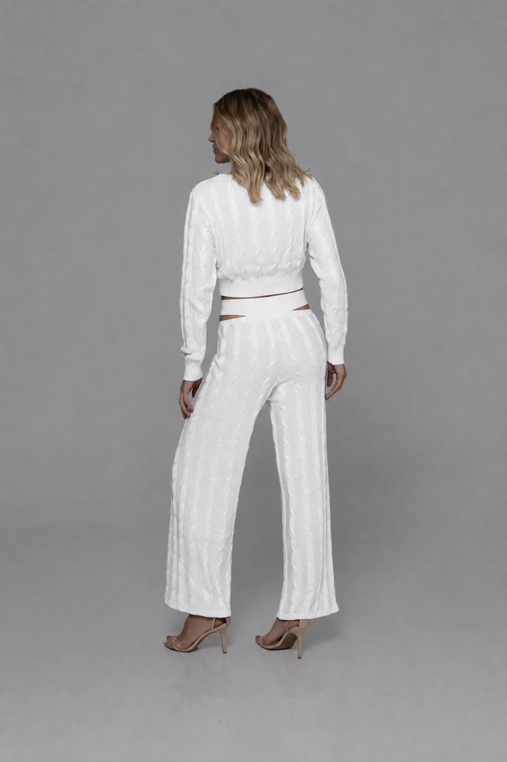 Casa Maly Bacana Set - White cable-knit crop top and matching trousers with distinctive waist cutouts, worn by a model on a Mediterranean balcony overlooking Portofino at sunset.