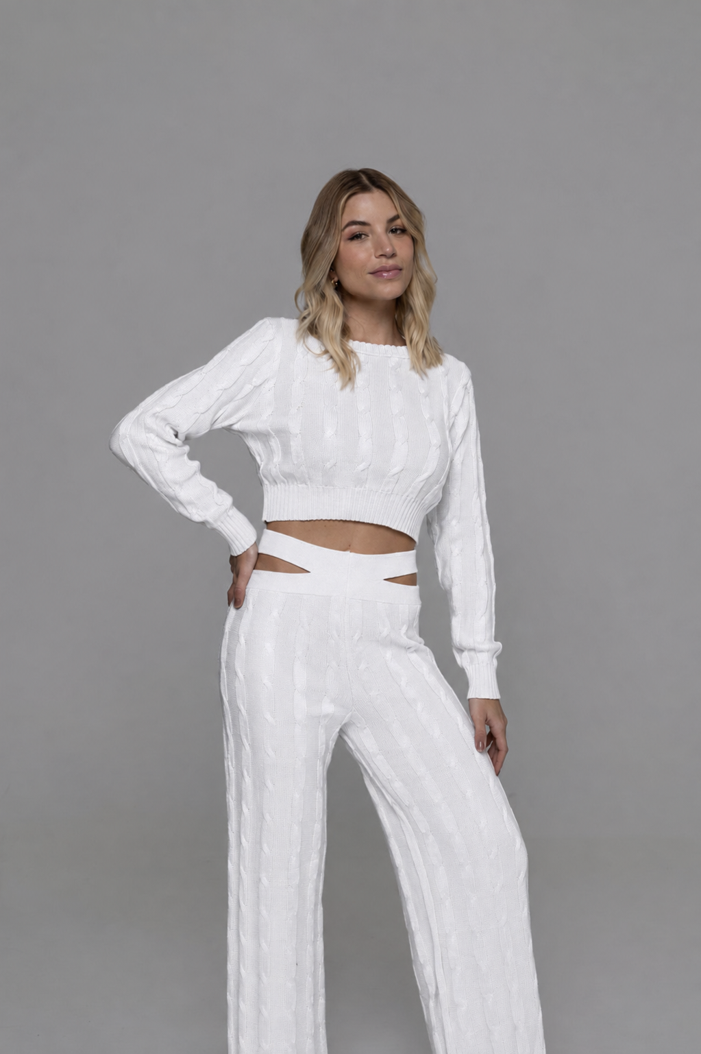 Casa Maly Bacana Set - White cable-knit crop top and matching trousers with distinctive waist cutouts, worn by a model on a Mediterranean balcony overlooking Portofino at sunset.