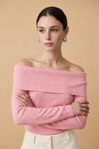 A woman wearing the Top Lecce off-the-shoulder knit top in pink, showcasing its elegant foldover neckline and long slim sleeves.