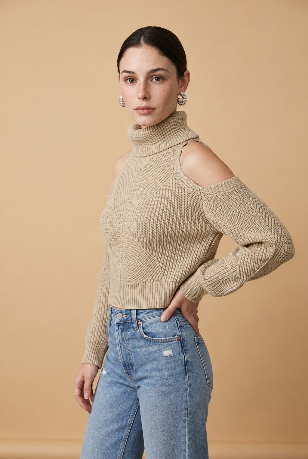 A woman wearing the Livigno cold-shoulder turtleneck sweater in beige, showing the textured chevron knit and high-neck silhouette.