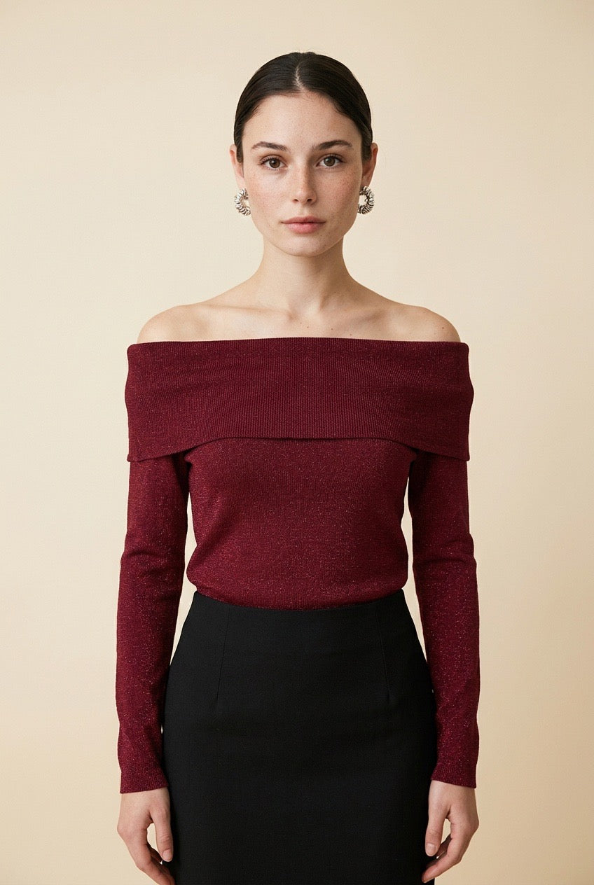 A woman wearing the Top Lecce off-the-shoulder knit top in burgundy shimmer, paired with a black tailored skirt.