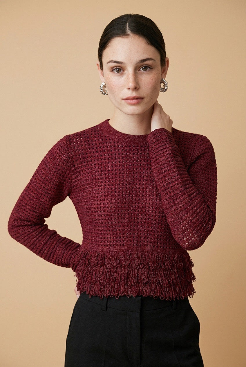 A woman wearing the Nerano long sleeve knit top in burgundy, featuring a crew neck, textured mesh stitch, and double-tiered fringe hem.