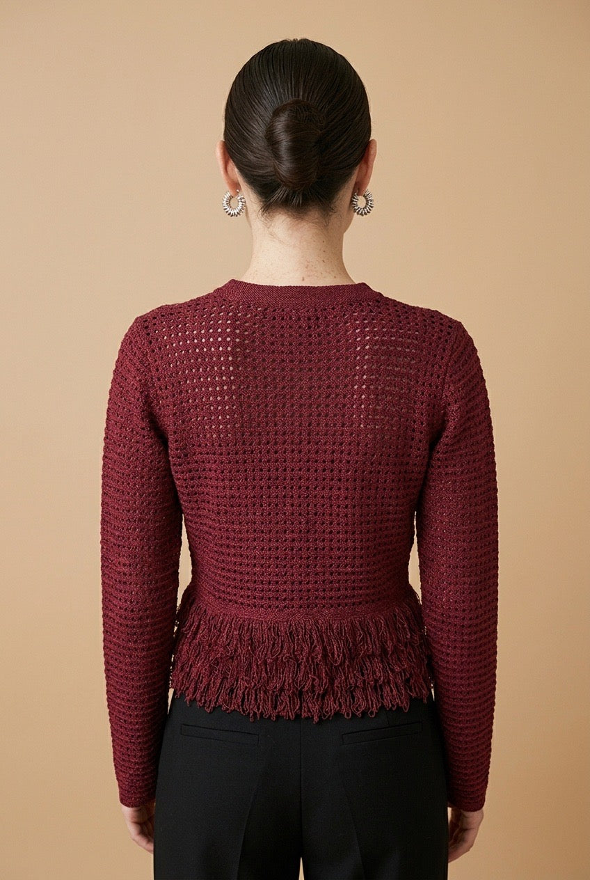 A woman wearing the Nerano long sleeve knit top in burgundy, featuring a crew neck, textured mesh stitch, and double-tiered fringe hem.