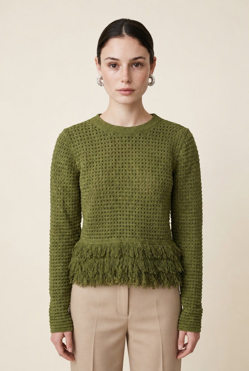 A woman wearing the Nerano long sleeve knit top in olive green, featuring a crew neck, textured mesh stitch, and double-tiered fringe hem.