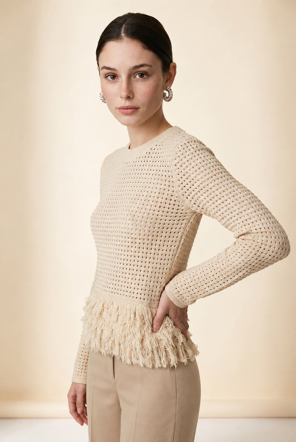 A woman wearing the Nerano long sleeve knit top in beige, featuring a crew neck, textured mesh stitch, and double-tiered fringe hem.