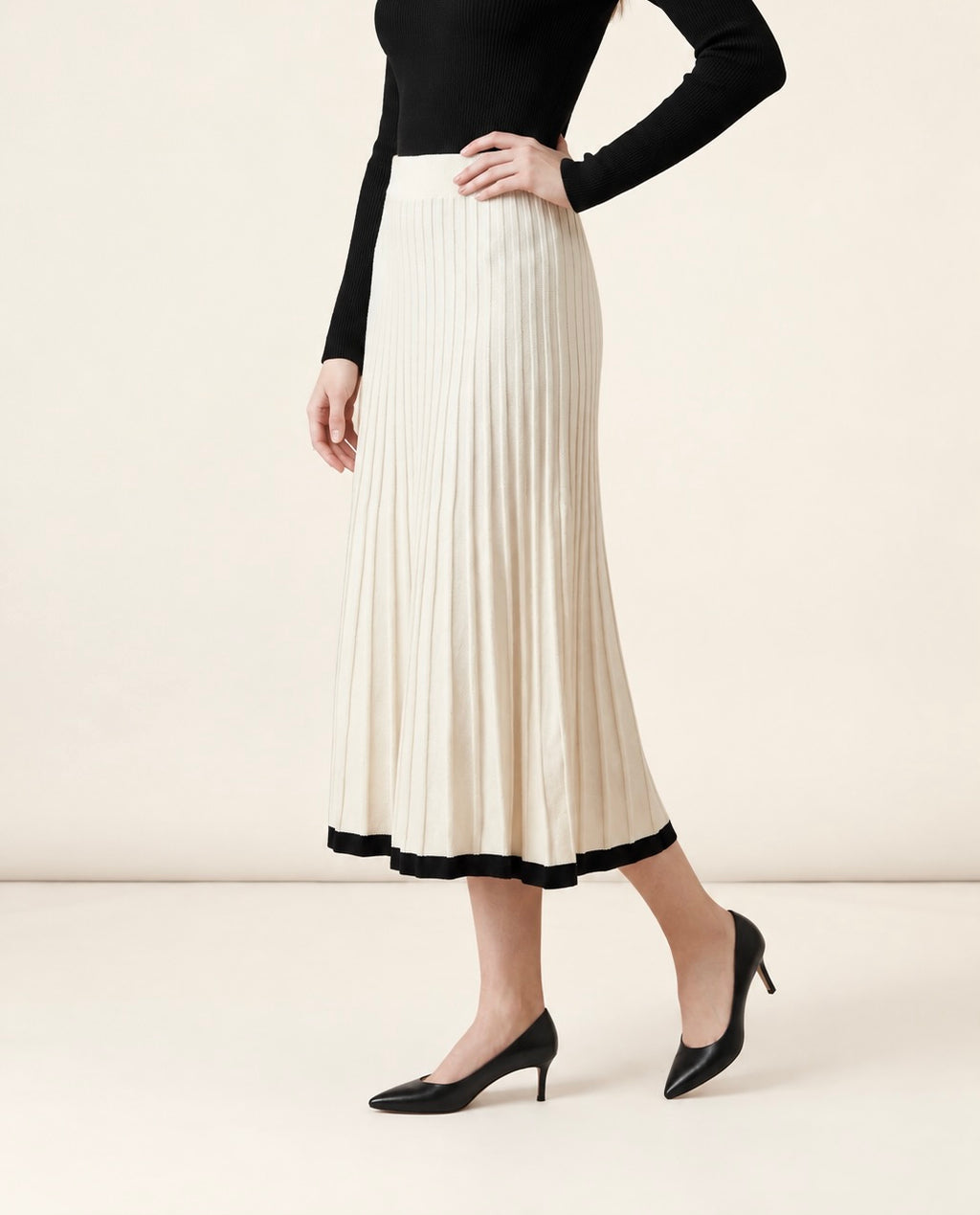 A woman wearing the Padova pleated knit midi skirt in off-white, highlighting its A-line flare and contrasting black hem line.