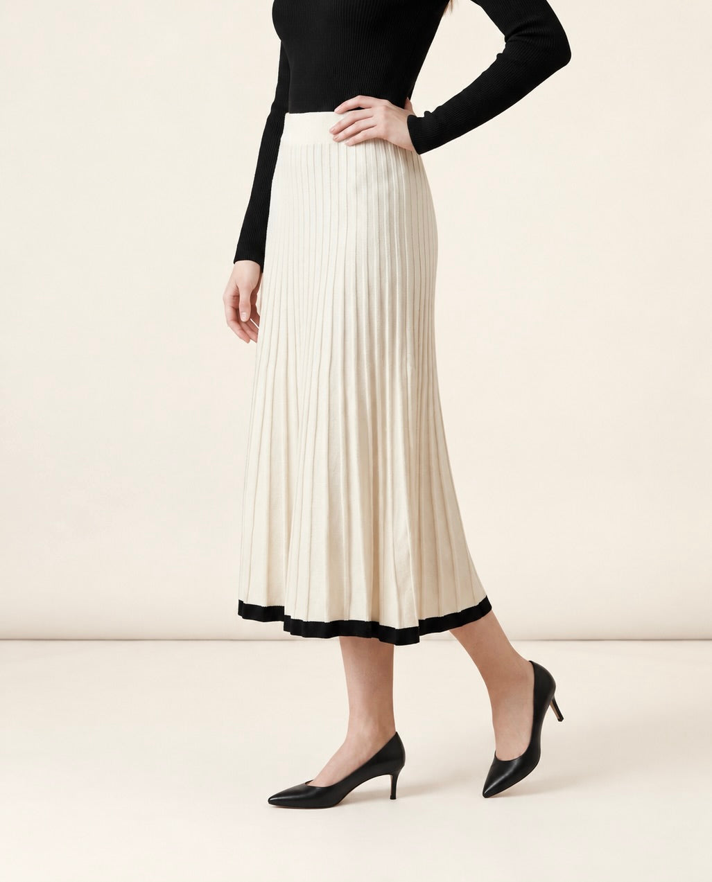 A woman wearing the Padova pleated knit midi skirt in off-white, highlighting its A-line flare and contrasting black hem line.