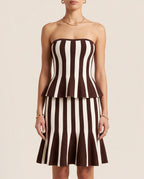 A model wearing a strapless bandeau-style dress with a peplum waist and flared skirt, featuring vertical brown and cream stripes, against a solid light beige background | Casa Maly