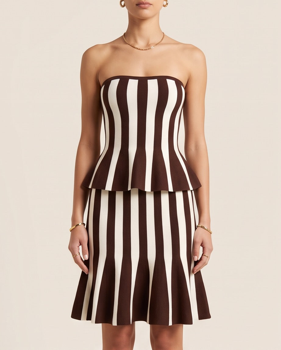 A model wearing a strapless bandeau-style dress with a peplum waist and flared skirt, featuring vertical brown and cream stripes, against a solid light beige background | Casa Maly