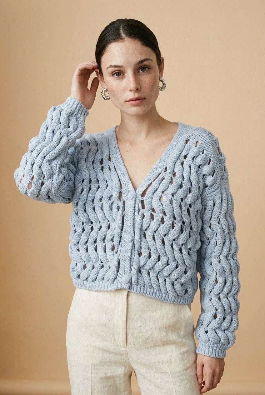 A woman wearing the Taormina open-knit cardigan in baby blue, featuring a button-down front and relaxed fit.