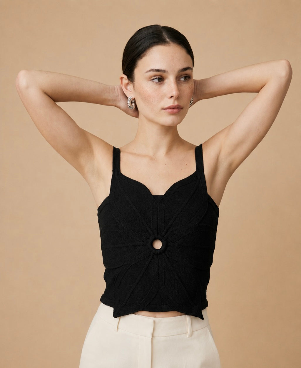 A young woman wearing a black sleeveless, cropped knit top with a central circular ring detail and a pointed asymmetric hem