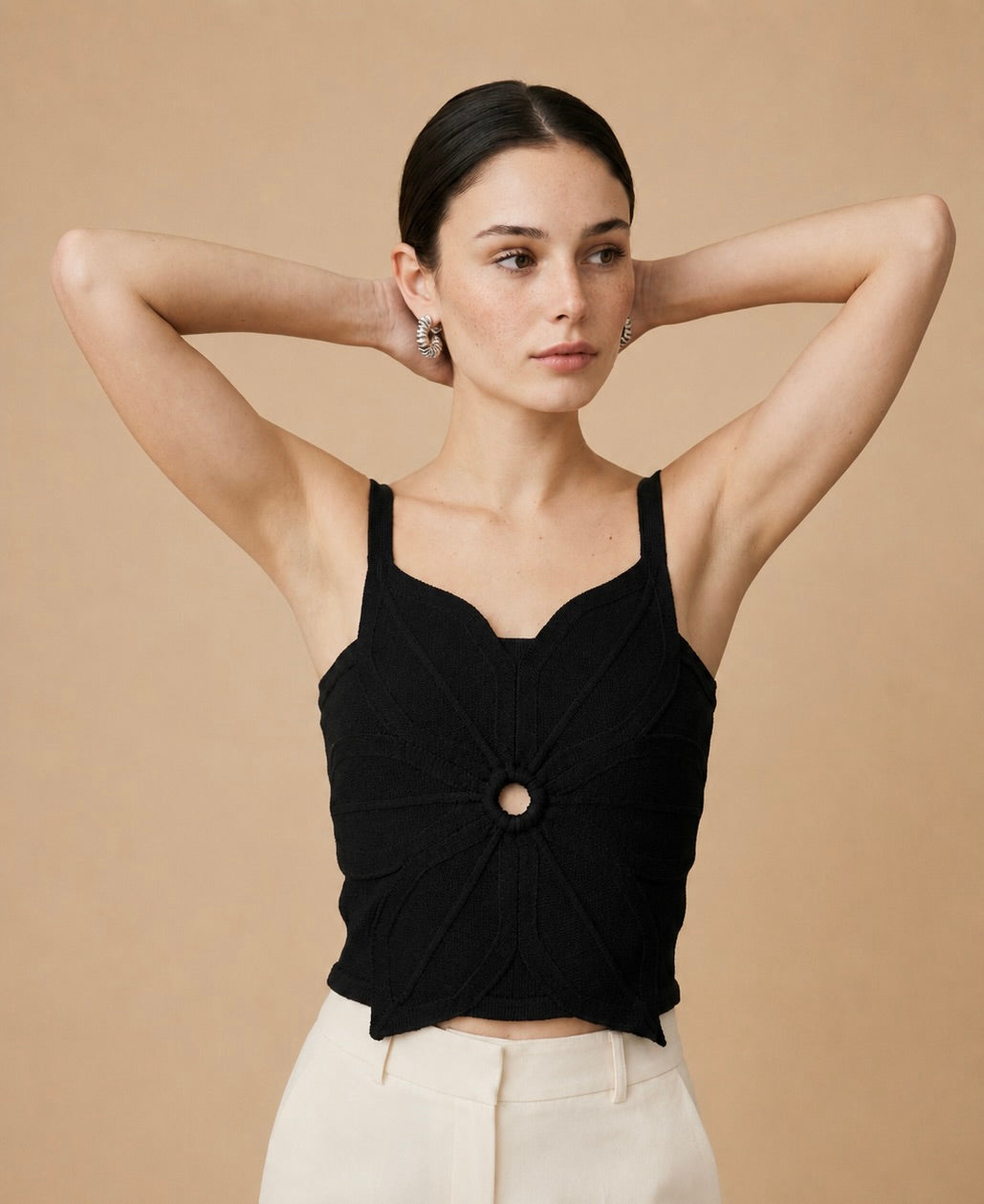 A young woman wearing a black sleeveless, cropped knit top with a central circular ring detail and a pointed asymmetric hem