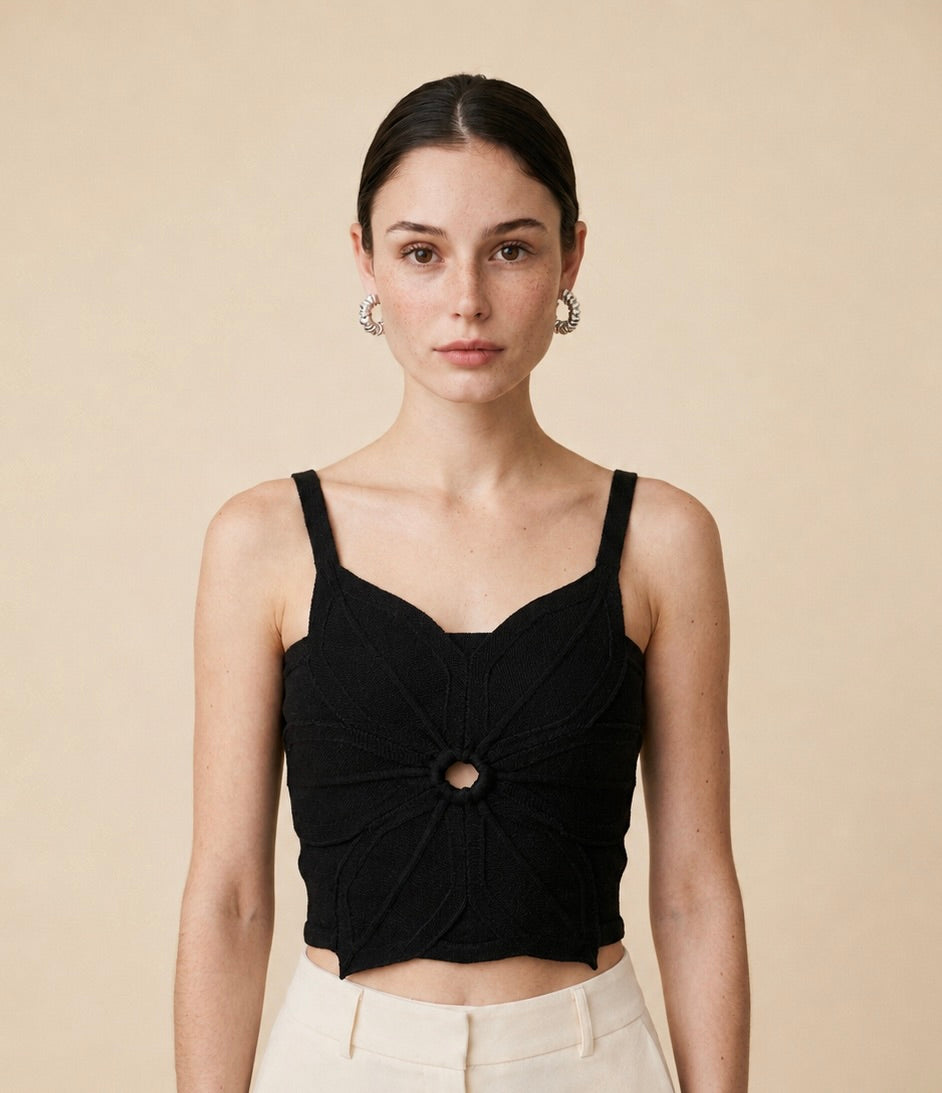 A young woman wearing a black sleeveless, cropped knit top with a central circular ring detail and a pointed asymmetric hem
